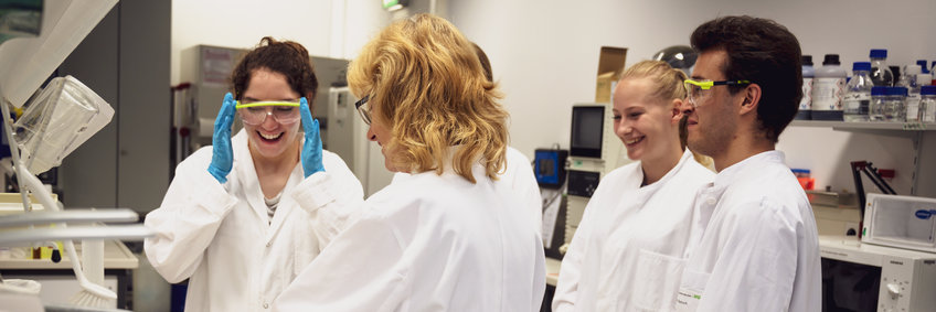 Laborszene mit vier lächelnden Personen im weißen Kittel, eine Frau fasst sich an die Schutzbrille.