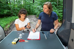 Two female scientists are standing at the open tailgate of a research vehicle in the forest. In front of them on a worktop are various working materials.