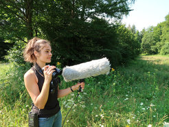 Female scientist standing with large microphone and binoculars in a meadow.